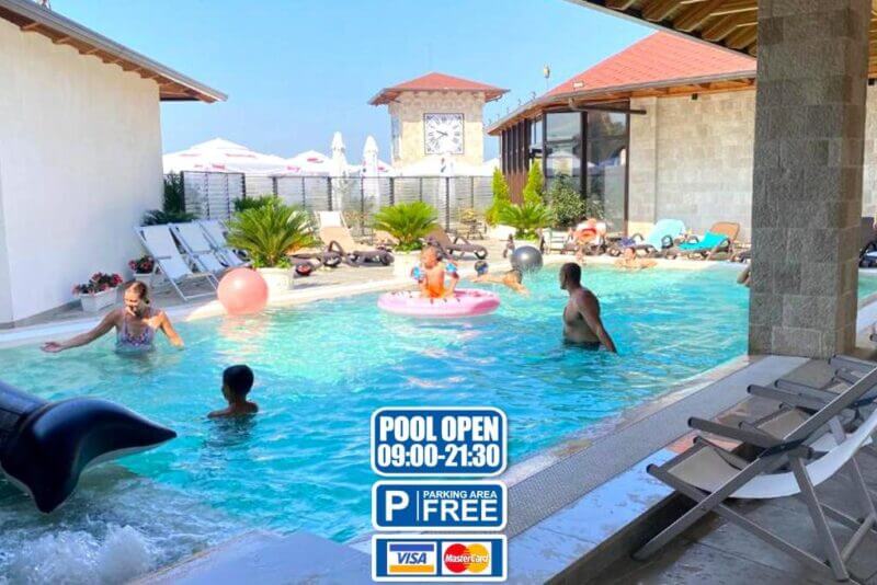 Indoor-outdoor pool area at Hotel Panorama with people swimming and relaxing; sign displays pool hours, free parking, and accepted payment methods (Visa, Mastercard).