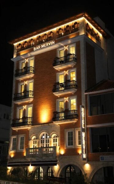 A multi-story brick hotel named Mervin Hotel and Bar Mervin is illuminated at night, with balconies on each floor and exterior lights highlighting the building’s facade.