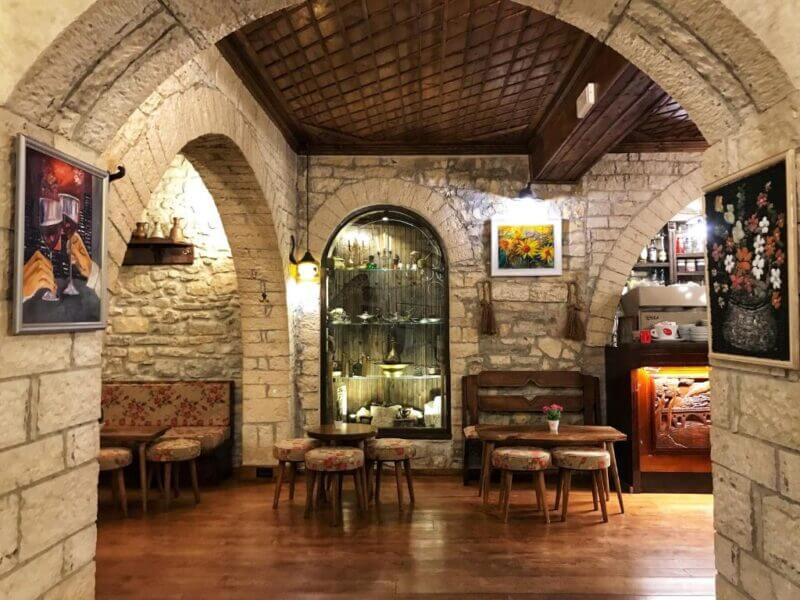 Cozy restaurant interior at Hotel Mangalemi with stone walls, wooden ceiling, small round tables with cushioned stools, wall art, and a display cabinet of decorative items.