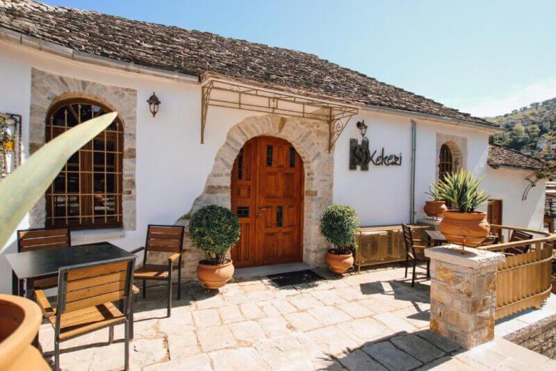 A stone building in Gjirokaster with a wooden door, arched windows, potted plants, and outdoor seating on a stone patio under a sunny sky—perfect for relaxing at the ss-kekezi hotel.
