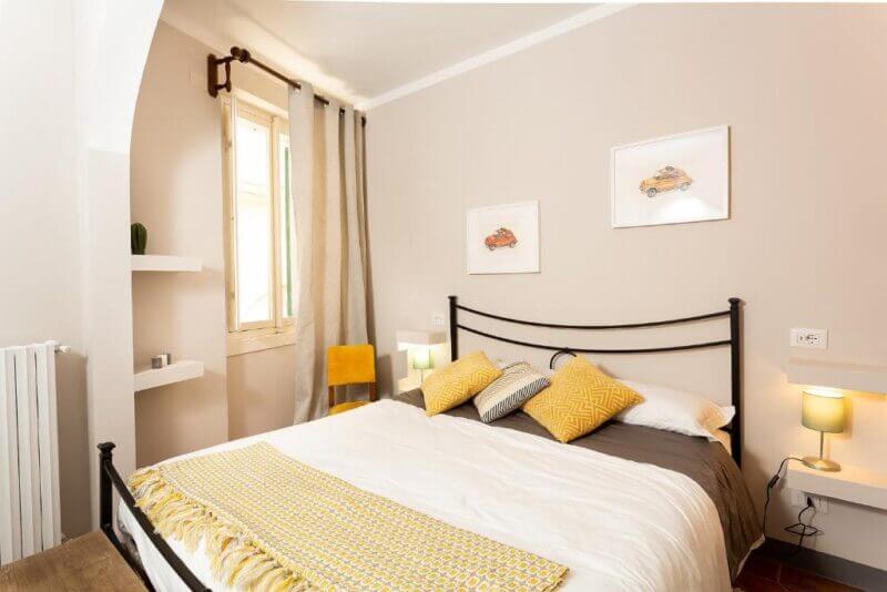 A neatly made bed with yellow and gray accents in a bright bedroom at Casa Dreosti, featuring two lamps, framed car prints, a window with curtains, and minimalist shelves—perfect for your stay in charming Cortona.