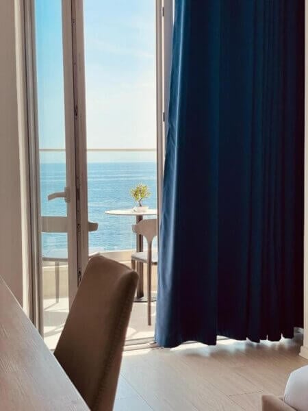 A room with light wood furniture, an open door leading to the Ionian Terrace balcony with a small table and plant, and stunning sea views under a clear Himare sky.