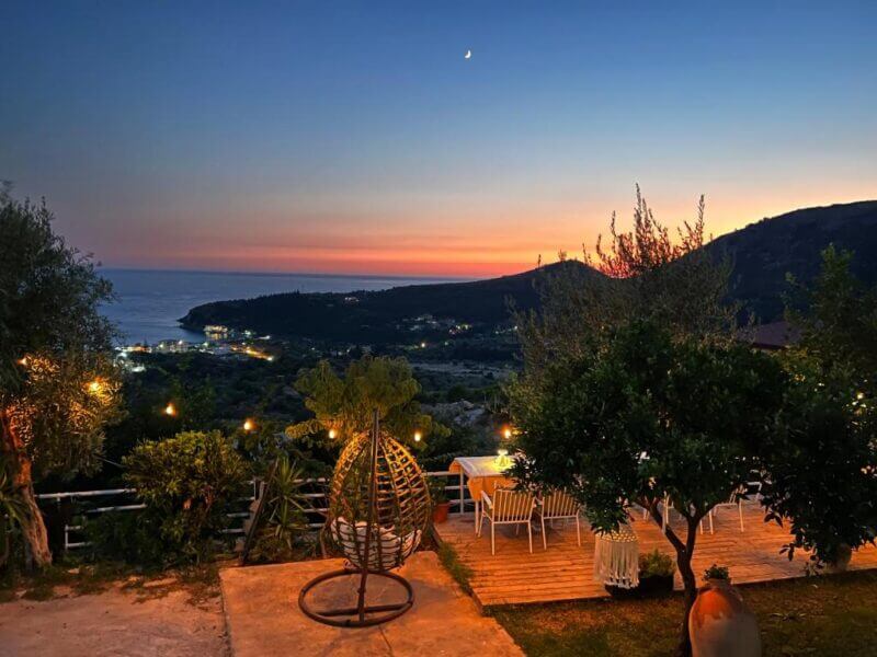 Outdoor seating area at Aphrodite Garden Rooms with string lights overlooks the coastal landscape of Himare at sunset, with hills, the sea, and a crescent moon visible in the sky.