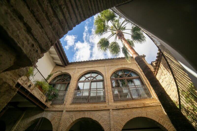 A view of the charming Patio del Posadero courtyard with arched windows, elegant columns, a tall palm tree, and blue sky with clouds above—a perfect Hotel Boutique escape.