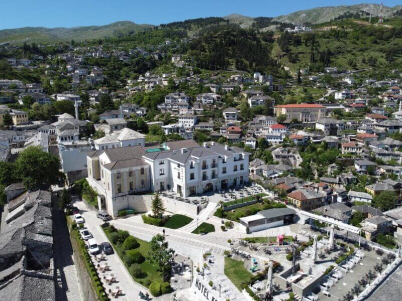 Aerial view of a hillside town with densely packed buildings, green hills in the background, and Hotel Fantasy Gjirokaster’s prominent white building with landscaped grounds in the foreground.