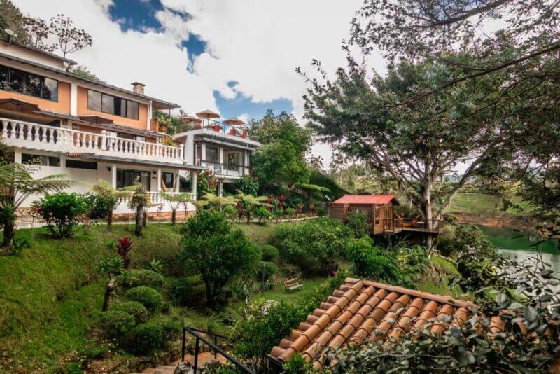 Two multi-story houses with balconies are surrounded by lush greenery, mature trees, and landscaped gardens near a small body of water in Guatape, offering a serene stay at Serendipity Hospedaje.