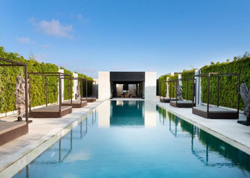 Rectangular outdoor swimming pool with cabanas and green hedges on each side, leading to a modern building under a clear blue sky at eqUILIBRIA Seminyak, a renowned Bali luxury resort.