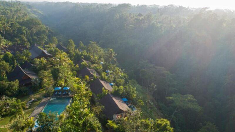 Aerial view of boutique resort in Ubud Valley, featuring thatched-roof villas, swimming pools, and lush tropical greenery nestled amid dense forest.