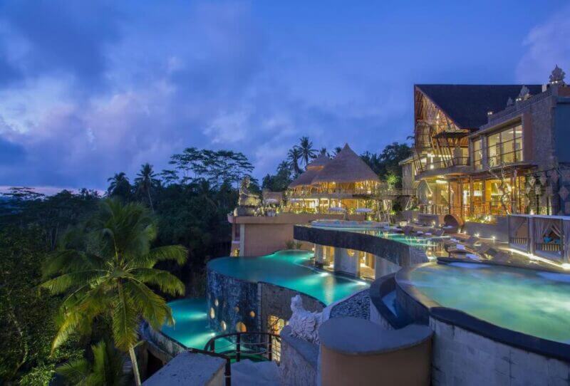 A luxury jungle resort at dusk with multi-level infinity pools, tropical palm trees, and illuminated buildings set on a lush Ubud hillside at the renowned Kayon Jungle Resort.