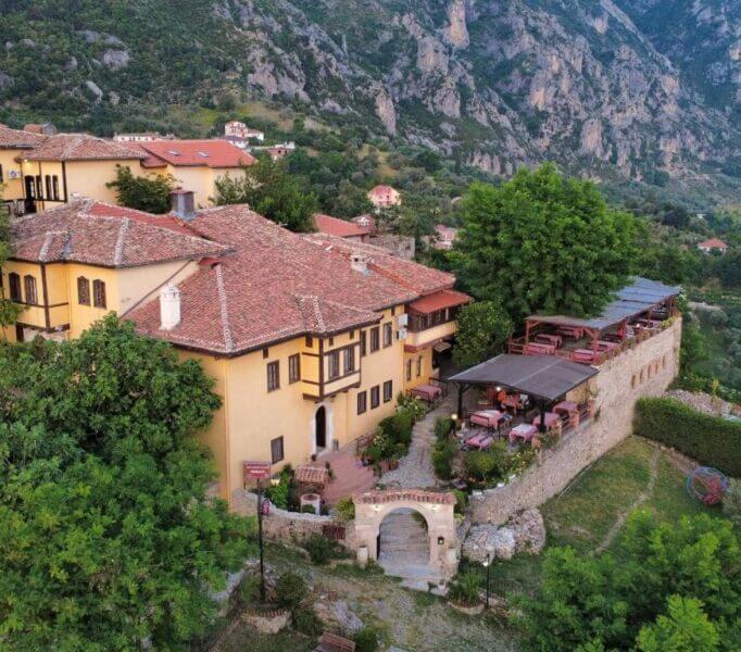 Aerial view of Rooms Merlika Kruje, a large yellow building with a red-tiled roof, nestled among trees and mountains, featuring an outdoor terrace with tables and chairs.
