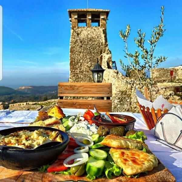 A table set with assorted Mediterranean dishes and vegetables is in the foreground, with the stone tower and blue sky near Rooms Emiliano Kruje in the background.