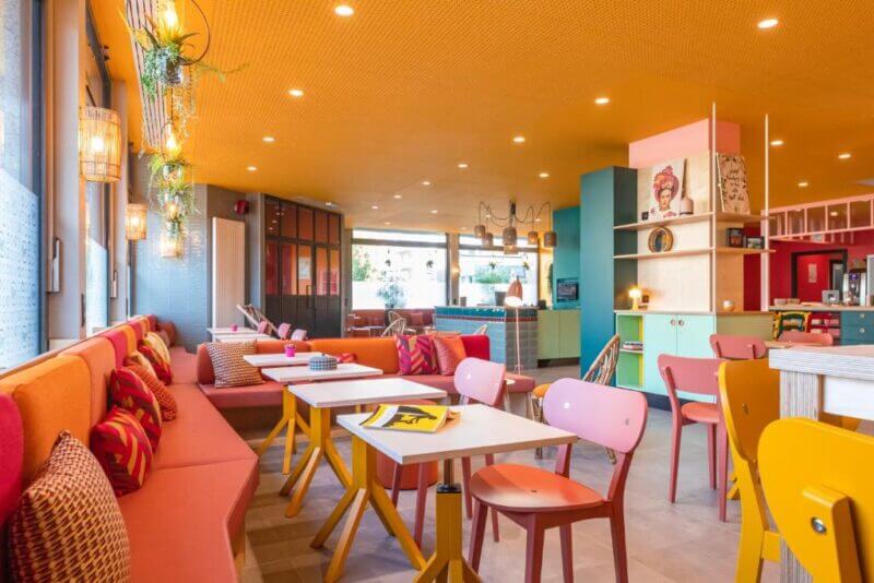 Colorful cafe interior by PAUL & PIA in Colmar with an orange ceiling, pink and yellow chairs, patterned cushions, wall plants, and large windows letting in natural light. Tables feature books and magazines for guests.