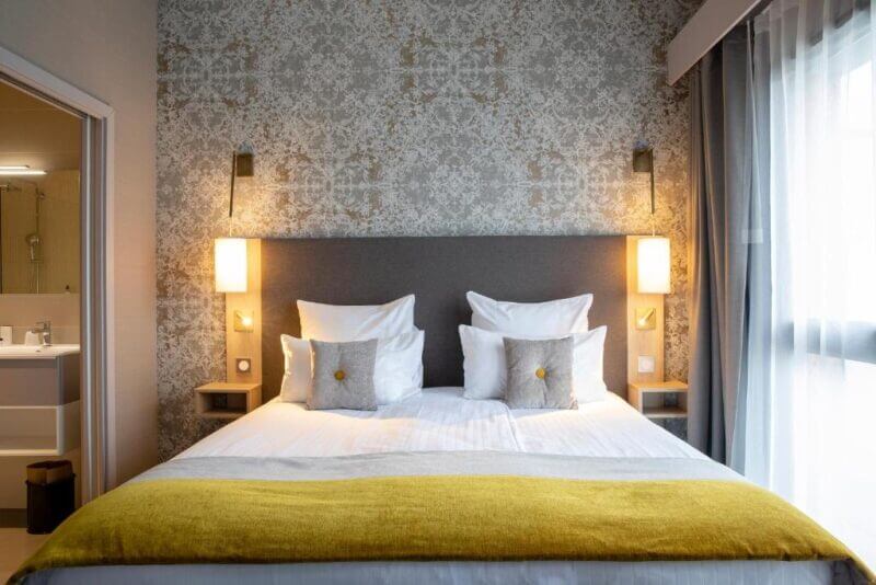Modern hotel room at James Boutique Hôtel in Colmar, featuring a double bed, two wall lamps, patterned wallpaper, yellow throw blanket, and an adjacent bathroom visible through an open door.