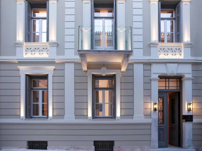 A beige, neoclassical building facade features large windows with decorative trim, a small balcony with glass railing, and illuminated wall lights, embodying the refined style of Kinglin Luxury Athens.