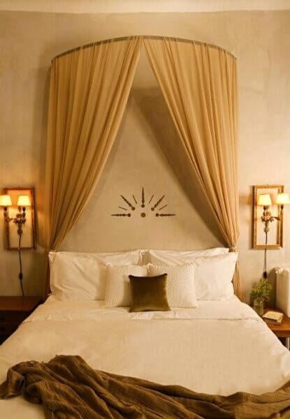 A neatly made bed with white linens, an olive green pillow, draped beige canopy, wall-mounted lamps, and a decorative wall accent above the headboard creates a serene retreat at Shila Athens boutique hotel.