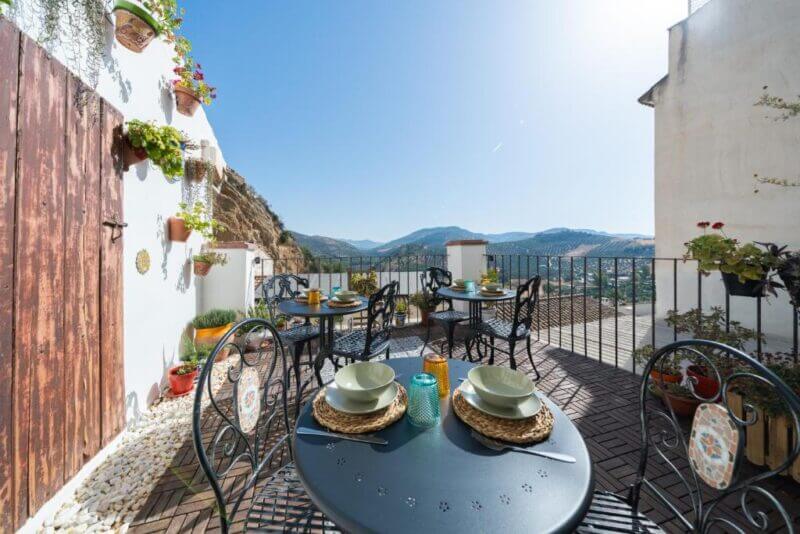Outdoor terrace at Villa Moana Iznajar with black metal tables set for a meal, surrounded by potted plants, overlooking mountains and a valley under a clear blue sky.