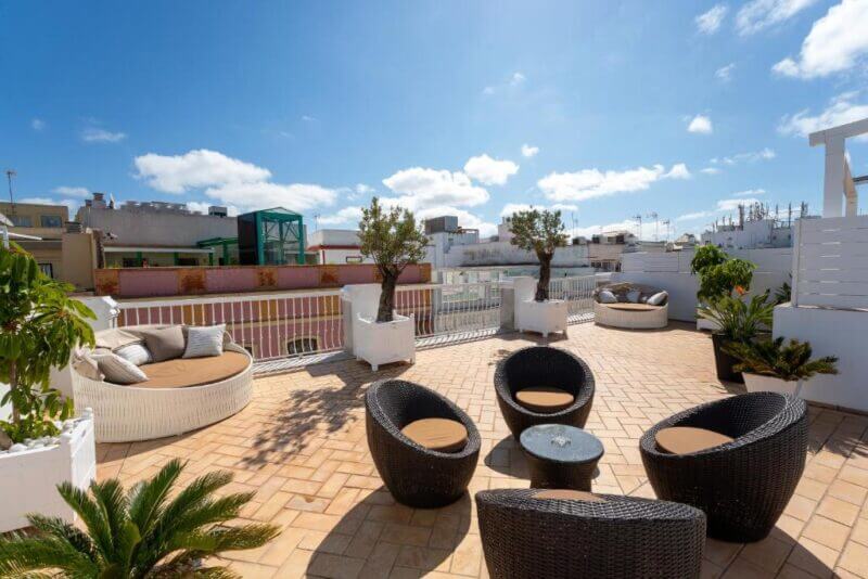 Rooftop terrace at Casa Palacio El Armador in Cadiz, featuring wicker chairs, round tables, potted plants, and views of surrounding buildings under a sunny, partly cloudy sky.