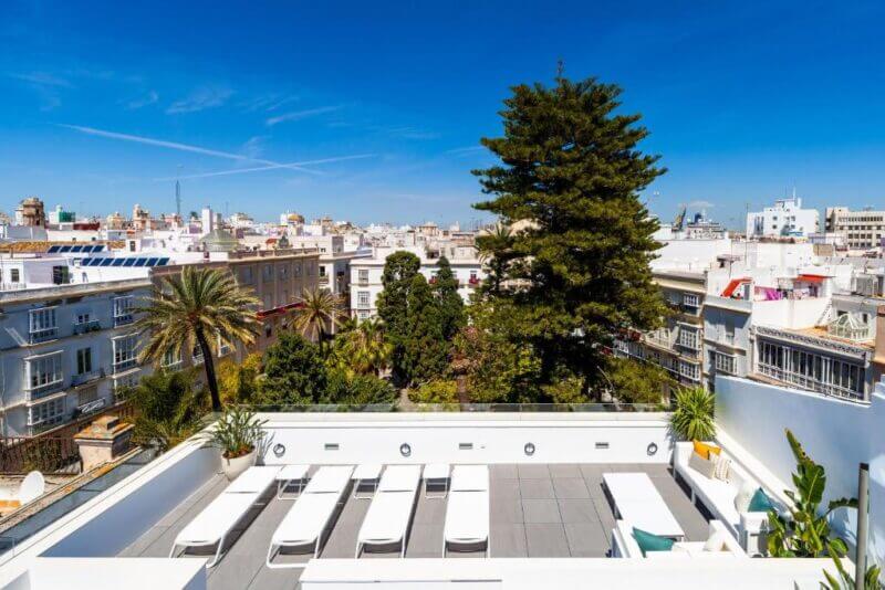 Rooftop terrace at Candelaria10 in Cadiz with white lounge chairs and potted plants, overlooking city buildings and a garden with tall trees under a clear blue sky.