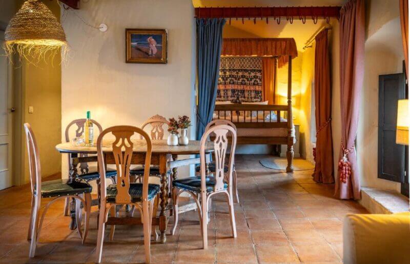 A cozy room in Ronda with a wooden dining table and chairs, a canopied bed in the background, tile flooring, warm lighting, and decorative curtains—perfect for enjoying Ronda Romántica or relaxing in charming lofts.