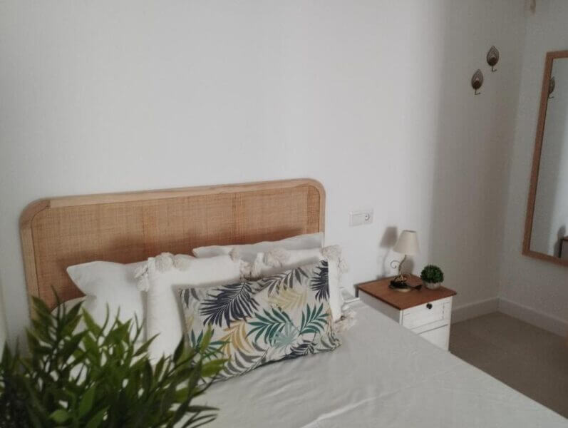 A neatly made bed with a wooden headboard, decorative pillows, a bedside table with a lamp and plant, and a wall mirror create a minimalistic bedroom retreat at Casa Eva Nerja.