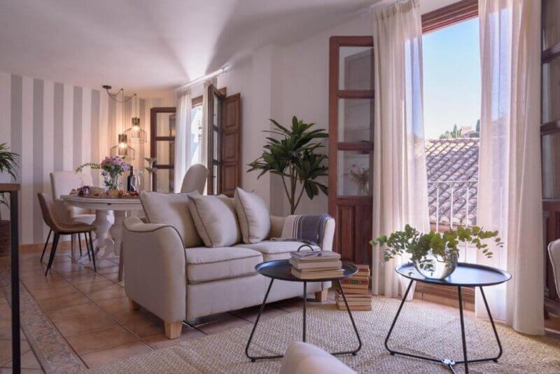 Bright living and dining area at Casa Boutique Granada with beige sofas, a round coffee table, potted plants, striped accent wall, and large windows with sheer curtains overlooking tiled rooftops.