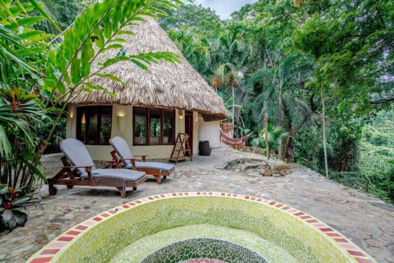 Thatched-roof bungalow at ONE Santuario boasts large windows, surrounded by lush tropical trees, with two lounge chairs and a mosaic-tiled hot tub on a stone patio—your perfect Palomino hotel escape.