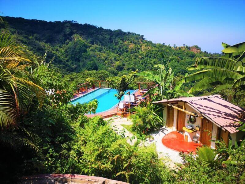 A small house in Casas Viejas with a red circular patio sits next to a rectangular pool, surrounded by dense tropical vegetation and hills under a clear blue sky.