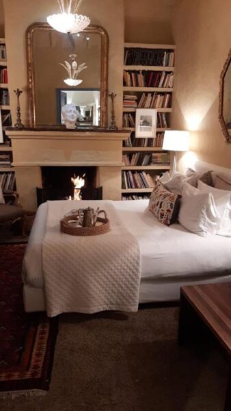 A cozy room at Candelaria House, a boutique hotel, features a bed facing a lit fireplace, bookshelves on either side, a mirror and decor above the mantel, plus a tray with cups and a teapot on the bed.