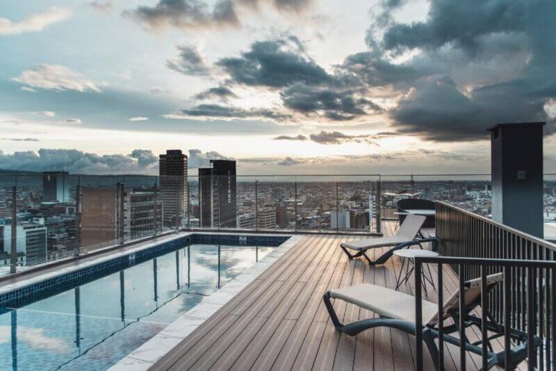 Rooftop pool with lounge chairs at Spotty Bogotá overlooks the city skyline and tall buildings of Bogotá Centro at sunset, with clouds drifting across the sky.
