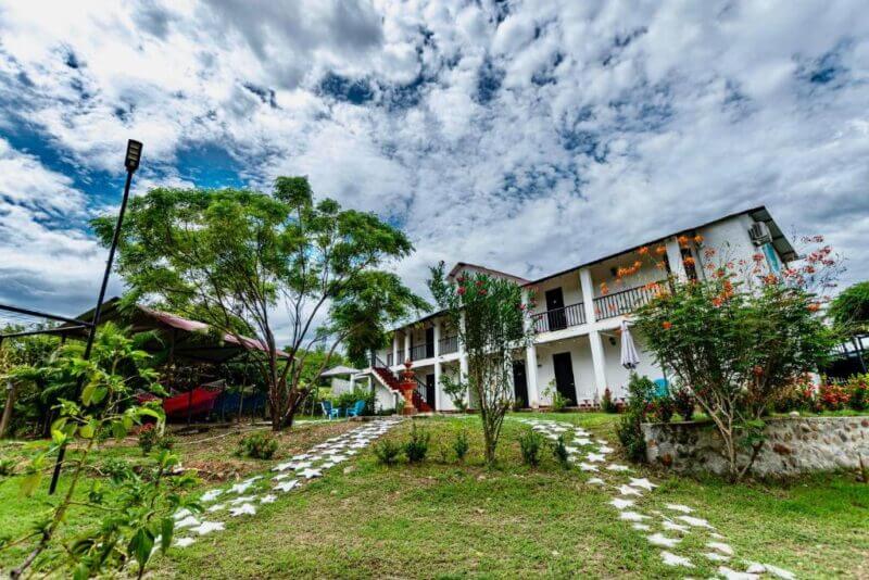 A two-story white building with balconies, part of Cosmos Tatacoa Hotel, sits in a landscaped yard with trees, bushes, and a stone path under a cloudy Tatacoa Desert sky.
