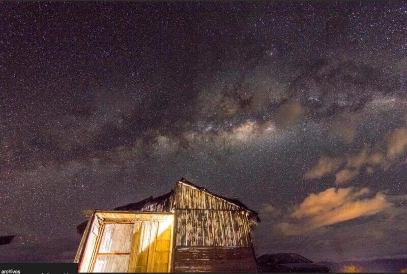 A rustic wooden hut sits illuminated under the clear night sky of Tatacoa, with stars and the Milky Way visible—an enchanting scene perfect for a glamping experience at Qji Glamping.
