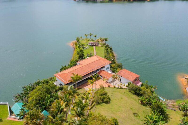 Aerial view of a house with a red-tiled roof, reminiscent of Guatape’s vibrant style, situated on a narrow peninsula and surrounded by a lake and lush greenery.