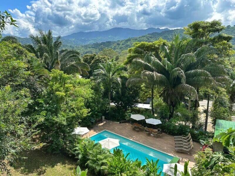 A rectangular outdoor pool at La Casablanca is surrounded by lush tropical trees, with lounge chairs and umbrellas, overlooking distant mountains near Tayrona under a partly cloudy sky.