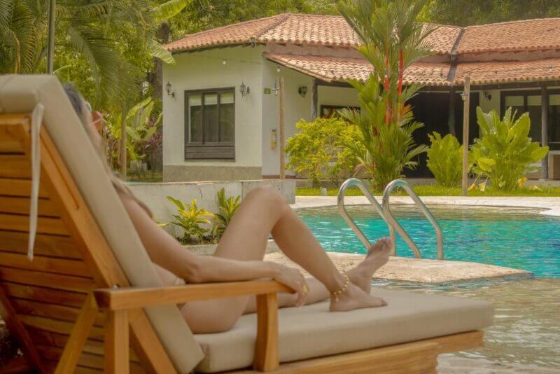 A person relaxes on a wooden lounge chair by a swimming pool at Hotel Tayrona, in front of a house with a tiled roof and lush greenery, enjoying the tranquil atmosphere near Kantawa Spa.