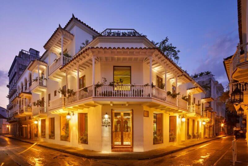 A well-lit, two-story white colonial-style building stands on a corner at dusk in Cartagena, featuring balconies with plants and large windows—reminiscent of the elegant Casona del Colegio—overlooking empty wet streets.