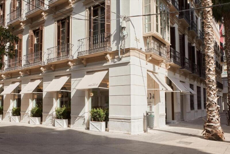 Street corner view of ICON Malabar, a beige multi-story building in Malaga with balconies and large windows, featuring awnings over the ground floor and potted plants along the sidewalk.