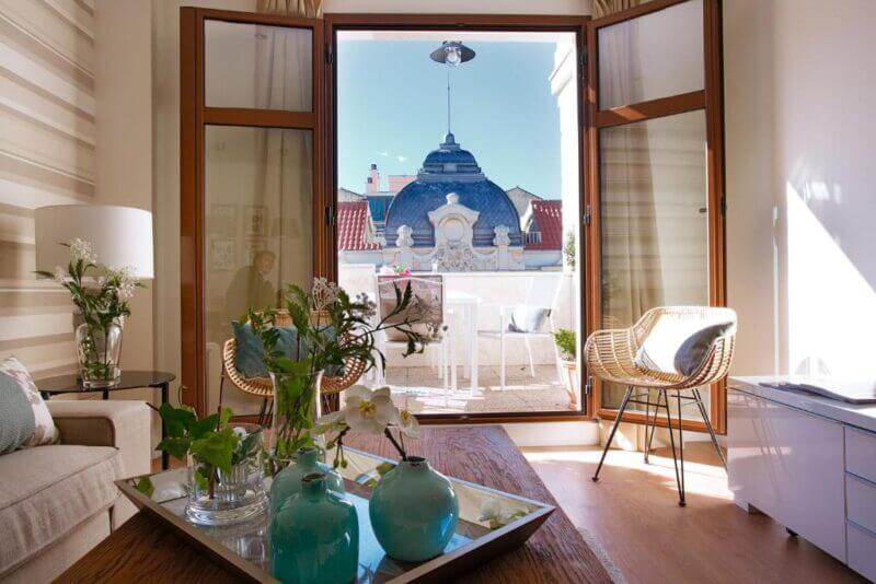 A bright living room with wooden floors opens to a balcony with chairs and a table, overlooking a building with a domed roof. Flowers and vases decorate a coffee table in this elegant space at iloftmalaga Hotel, your ideal Malaga hotel retreat.