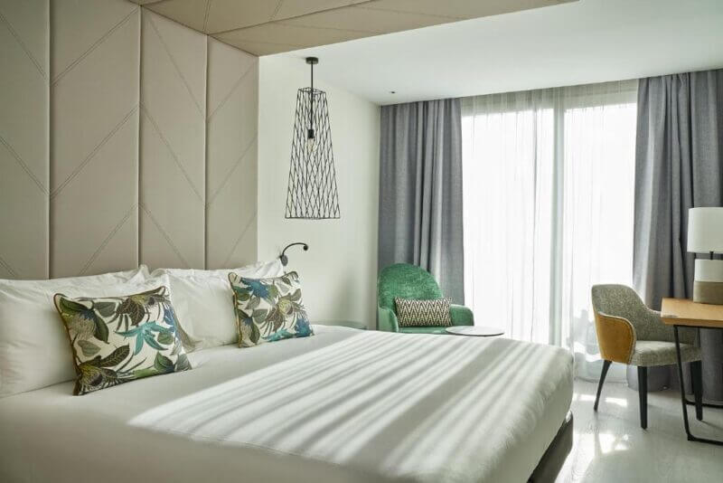 Modern Vincci Larios Diez hotel room in Malaga featuring a large bed, decorative pillows, green armchair, desk with chair, and floor-to-ceiling window with sheer curtains letting in natural light.
