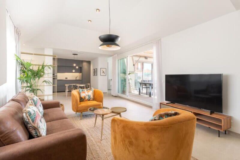 Modern living room at Be Mate Hotel Malaga, featuring a brown sofa, two mustard yellow armchairs, a large flat-screen TV on a wooden stand, and an open kitchen in the background.