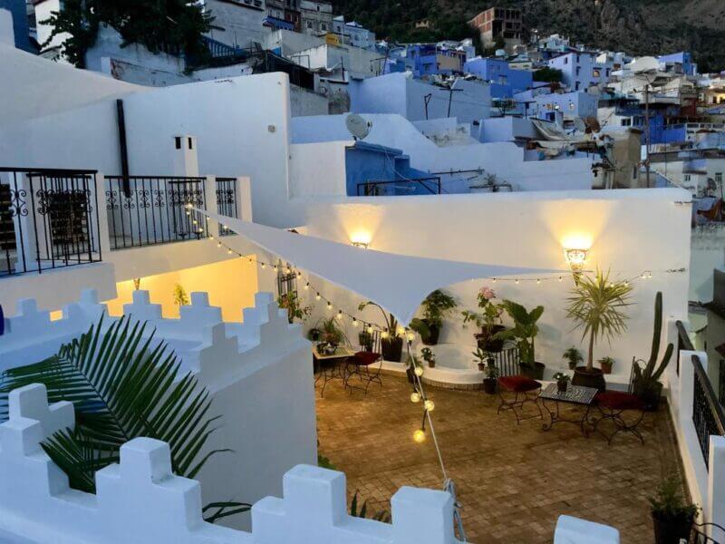 Rooftop terrace at Dar Hannan with white walls, potted plants, and string lights features outdoor seating overlooking a Chefchaouen hillside of blue and white buildings at dusk.