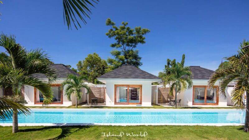 Three white villas with dark roofs face a long rectangular swimming pool at Island World Bohol, surrounded by palm trees and greenery under a clear blue sky.