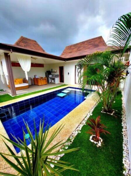 Small outdoor pool with blue tiles, surrounded by plants and white stone, next to a patio with seating, kitchen, and wooden roof—this Bali Villas-inspired haven in Panglao, Bohol rests under a cloudy sky.