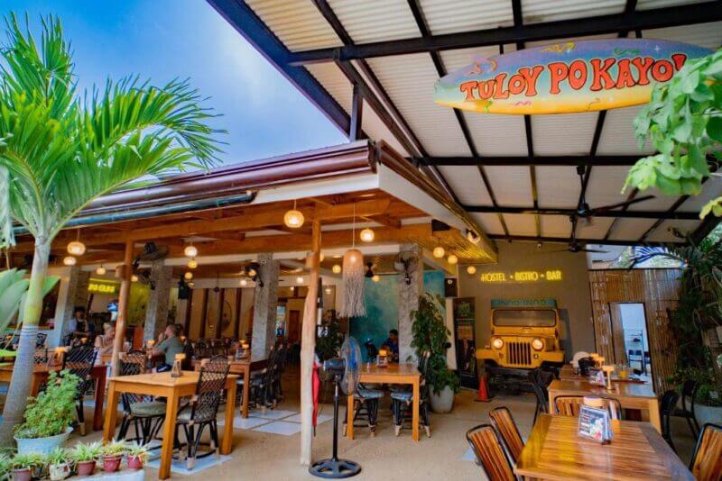 Outdoor seating area of Guni Guni restaurant in Puerto Princesa, with wooden tables, chairs, hanging lights, potted plants, and a sign reading Tuloy Po Kayo. A jeep-themed bar is visible inside.