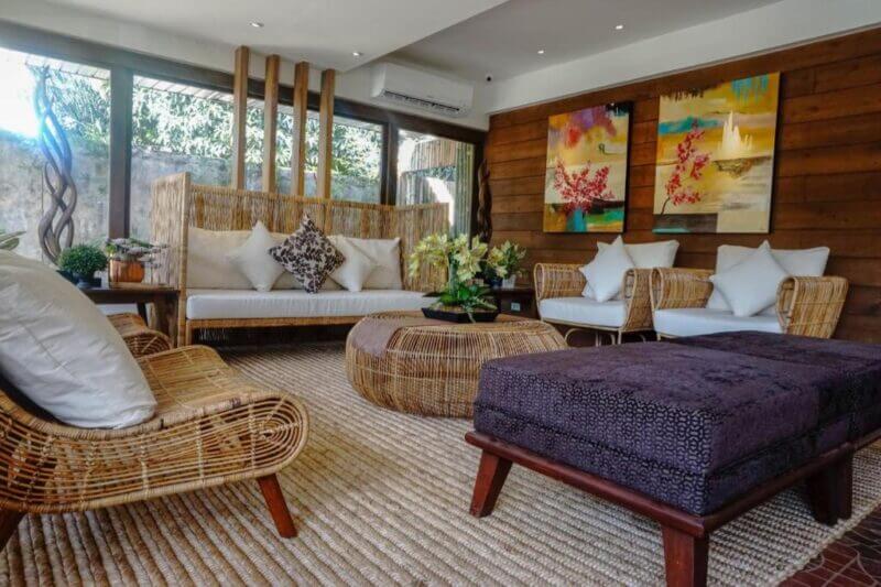 A modern living room at Balai Puerto Princesa features wicker furniture, white cushions, a round coffee table, wall art, indoor plants, and large windows that let in an abundance of natural light.