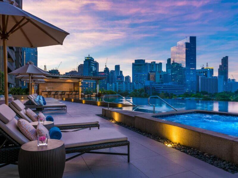 Poolside lounge chairs and a hot tub at Discovery Primea Manila overlook a city skyline at sunset, with drinks placed on a side table and tall buildings in the background.