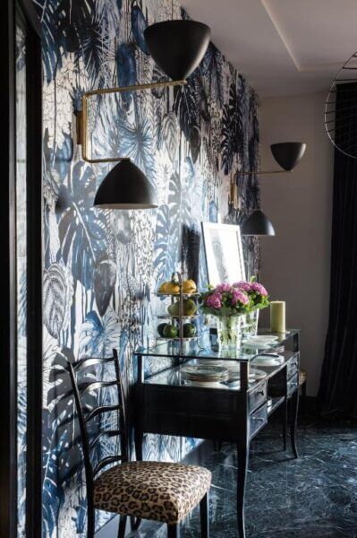 A glass console table with dishes, flowers, and a fruit stand sits against the blue botanical Velona's Jungle wallpaper; black sconce lights and a chair with a leopard print seat complete this stylish Florence-inspired setting.