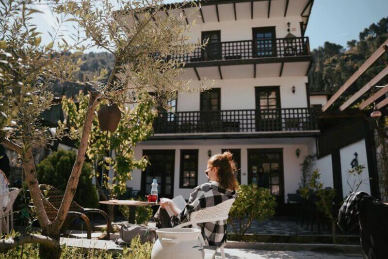 A woman sits at an outdoor table reading in front of Maya Hostel Berat, a white, two-story building with balconies, surrounded by greenery on a sunny day.