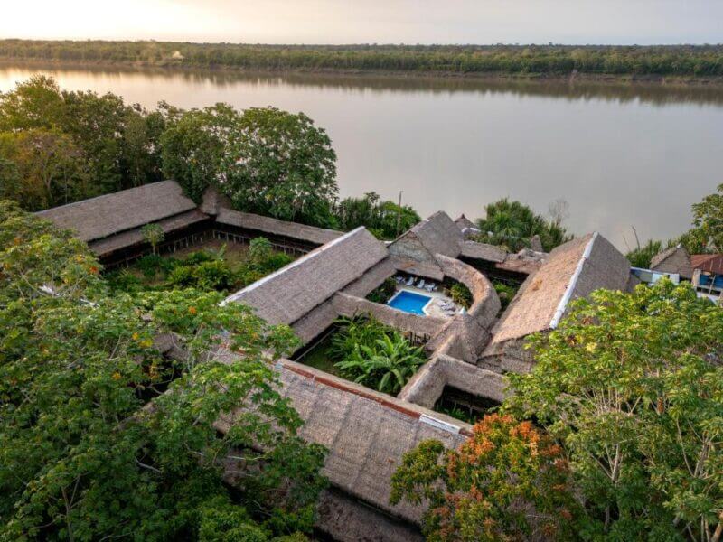 Aerial view of Amazon River Lodge with a thatched-roof, surrounded by trees and Heliconia blooms, featuring a central pool beside the wide Amazon River under a cloudy sky.