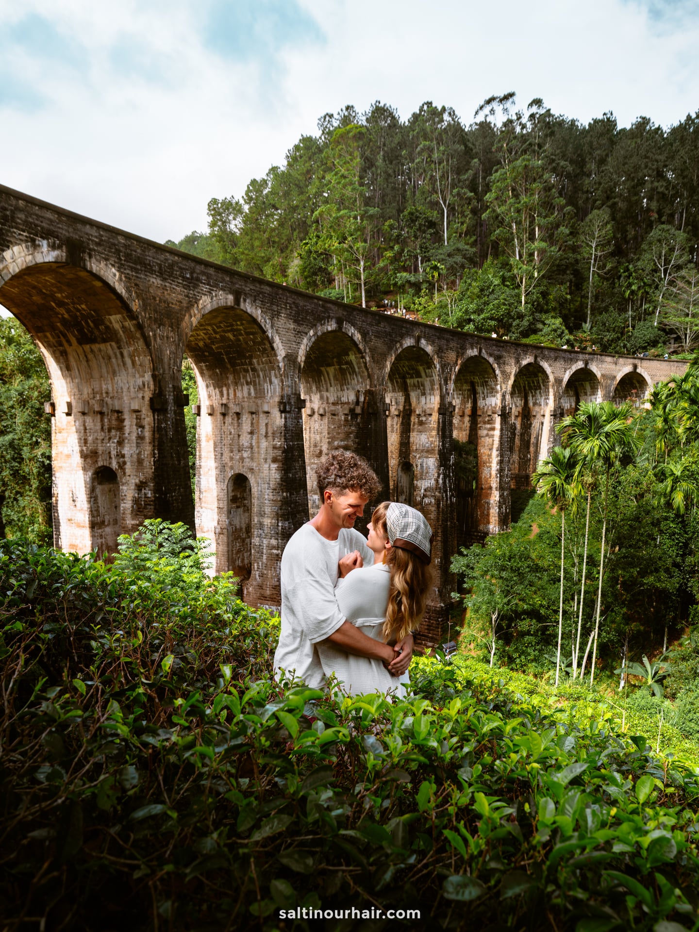 A couple stands embracing in front of a large stone railway viaduct called the Nine Arch bridge in Ella, Sri Lanka, surrounded by lush greenery and tall trees.