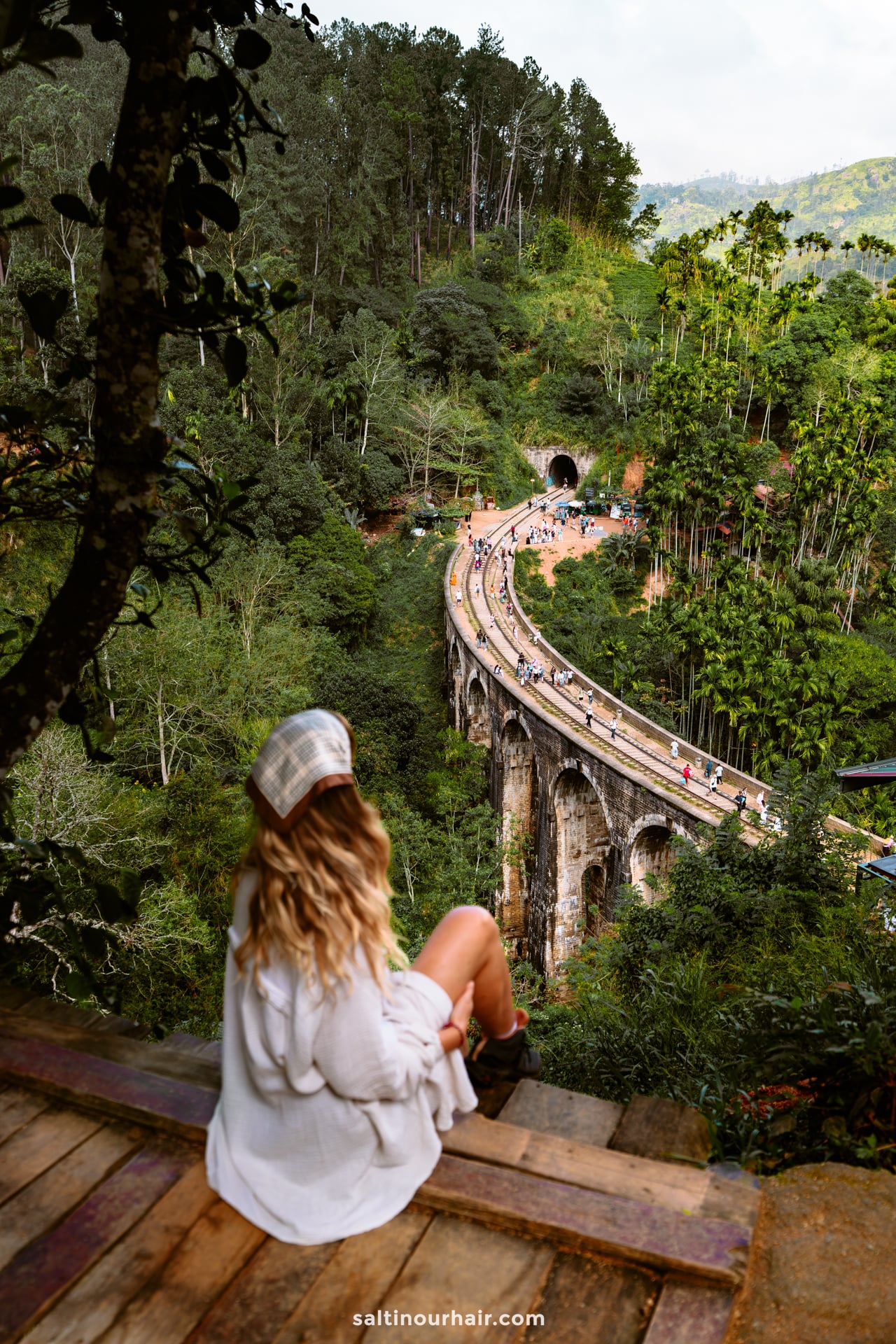 A woman in a white outfit sits on a wooden ledge overlooking the famous curved stone railway nine arch bridge in Ella, Sri Lanka, surrounded by lush green forest.